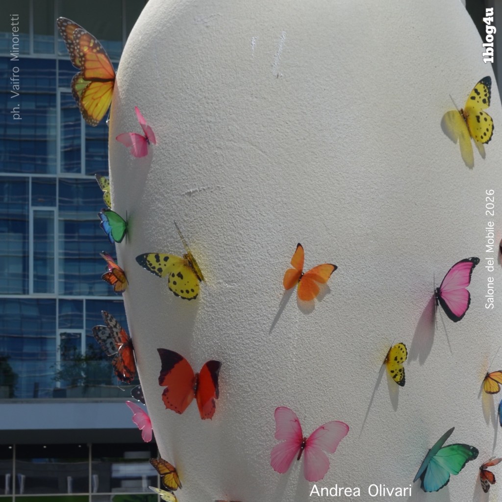 A sculpture with a smooth white surface adorned with colorful artificial butterflies in various shades and patterns, set against a modern building background.