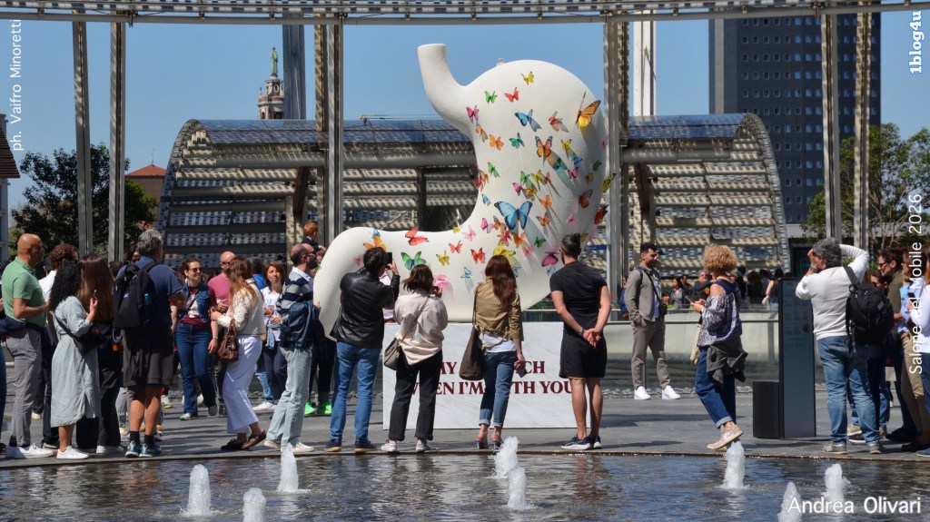 A large white sculpture of an elephant adorned with colorful butterflies, surrounded by a crowd of people admiring it, set in a public space with modern architecture and a fountain.