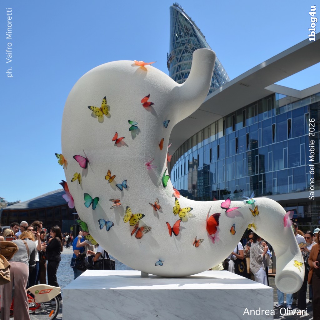 A large, white sculptural piece resembling a stomach, decorated with colorful butterfly motifs, set against a modern architectural backdrop during an outdoor event.