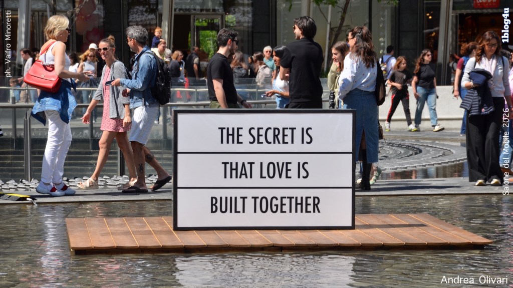 A group of people walking in a busy urban area near a reflective pool. In the foreground, a large sign displays the message 'THE SECRET IS THAT LOVE IS BUILT TOGETHER.'